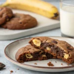 Freshly baked chocolate chip cookies with banana chunks, served with a glass of milk on a rustic table.