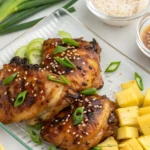 A top-down view of grilled Hawaiian BBQ chicken thighs, garnished with fresh green onions and sesame seeds, served alongside chunks of fresh pineapple and a drizzle of coconut glaze.