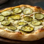 Side view of a freshly baked pickle pie pizza with melted cheese and sliced pickles on a wooden serving board.