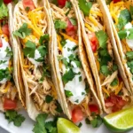 A plate of chicken tacos garnished with shredded cheese, diced tomatoes, sour cream, and fresh cilantro.