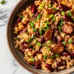 A bowl of red beans and rice with sausage and chopped green onions on a marble countertop.