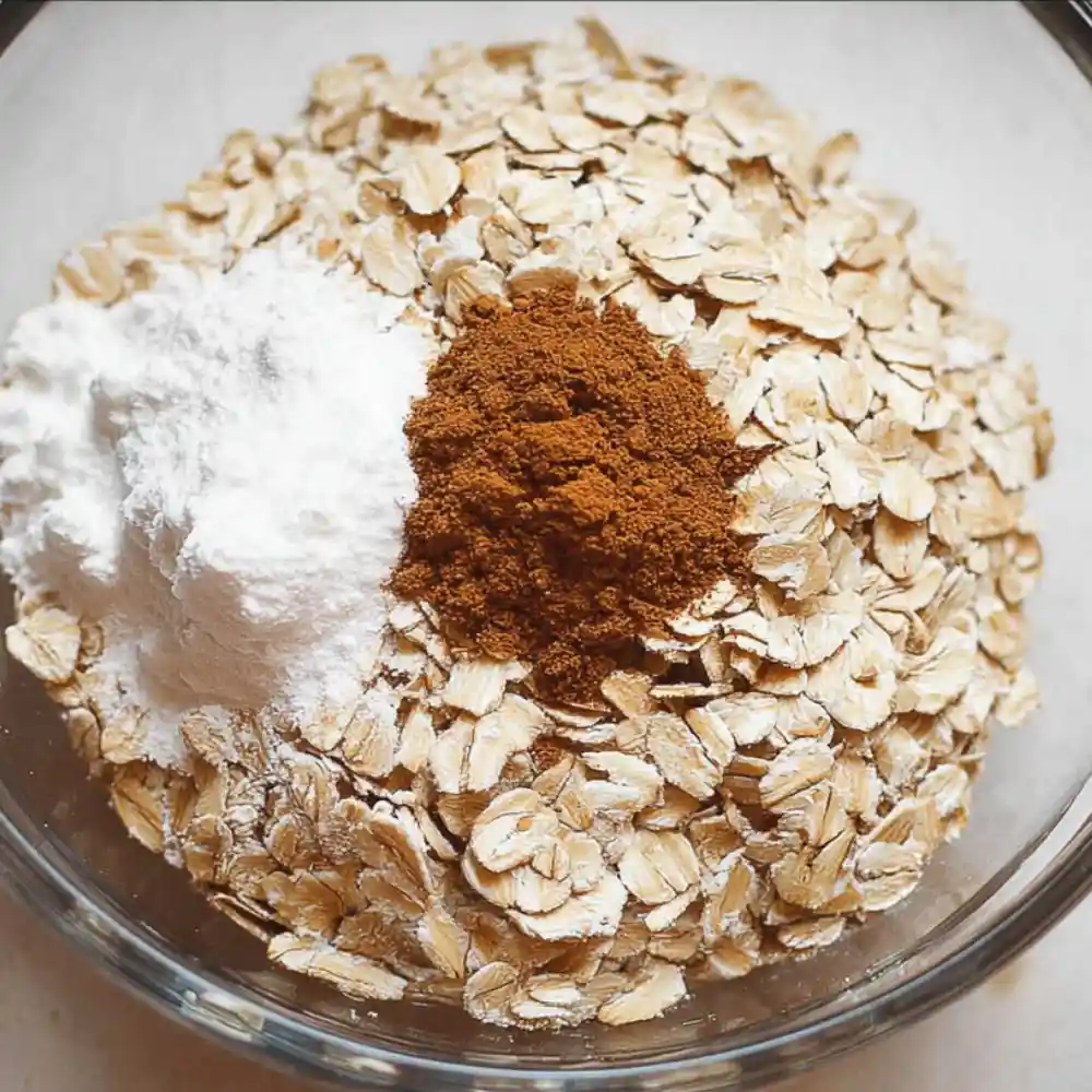 Rolled oats, baking powder, and cinnamon in a glass bowl for baked oatmeal preparation.