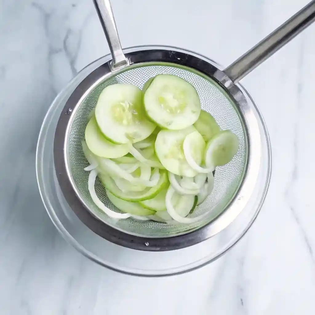 Sliced cucumbers and onions draining in a metal strainer over a glass bowl.