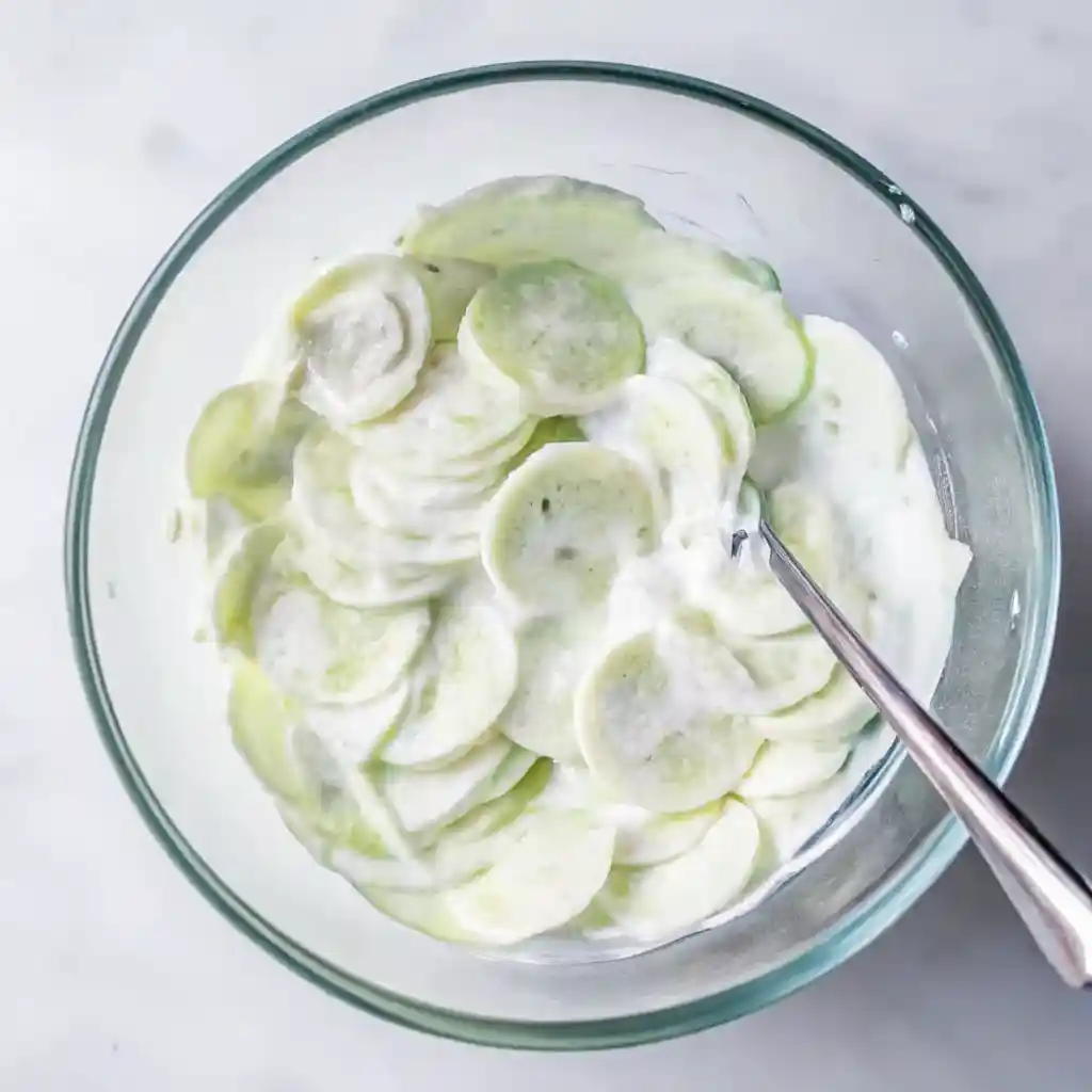 Thinly sliced cucumbers in creamy dressing in a glass bowl with a metal spoon.