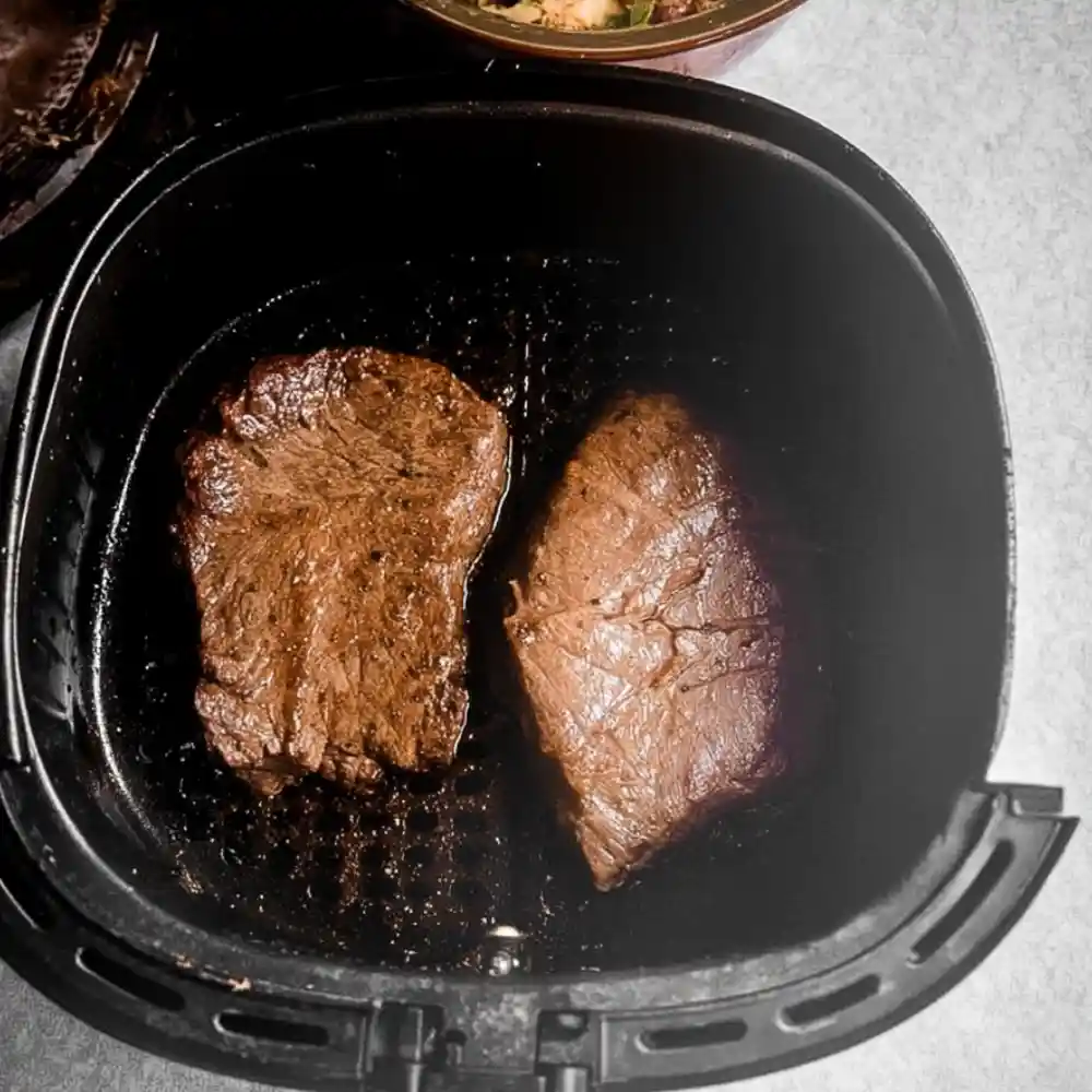 Two seasoned steaks cooking in an air fryer basket on a kitchen counter.