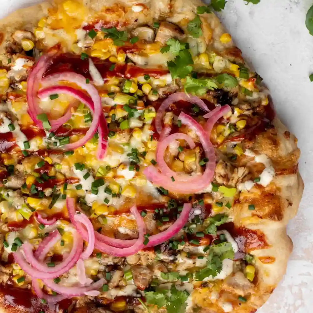 Top view of grilled BBQ chicken pizza with corn, cheese, ranch drizzle, cilantro, and pickled red onions on a crispy crust.