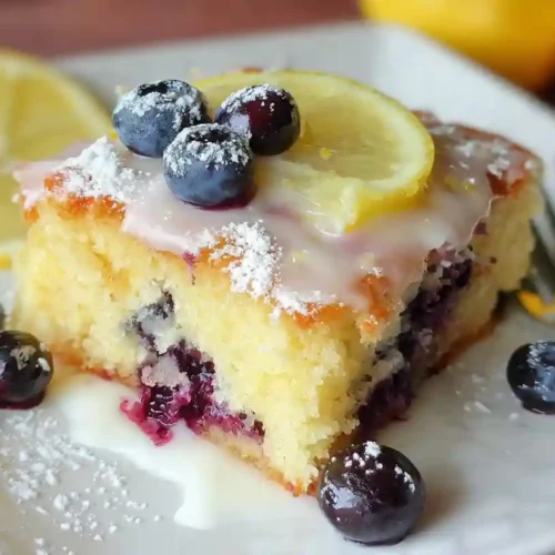 Moist lemon blueberry sheet cake topped with lemon glaze, fresh blueberries, and a lemon slice on a white plate.