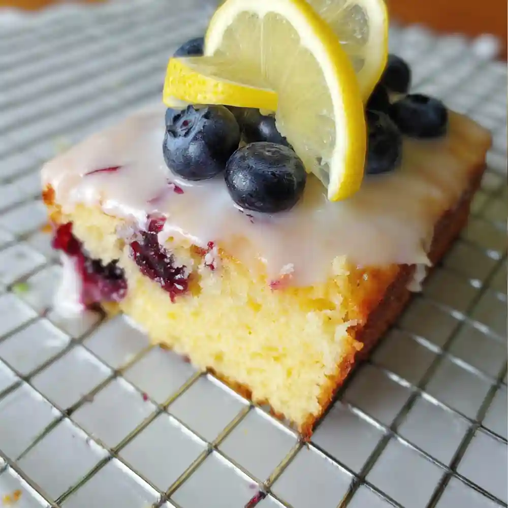 Lemon blueberry sheet cake slice with lemon glaze, topped with blueberries and lemon slices on a cooling rack.