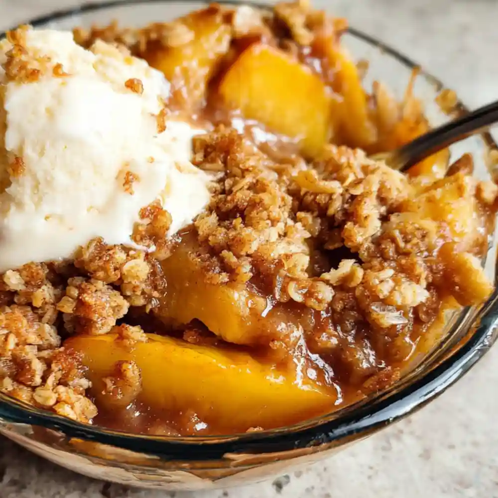 Warm peach crisp topped with vanilla ice cream in a glass bowl.