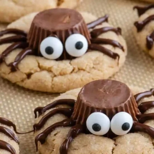 Close-up of peanut butter spider cookies with Reese’s cups, chocolate legs, and candy eyes on a baking sheet.