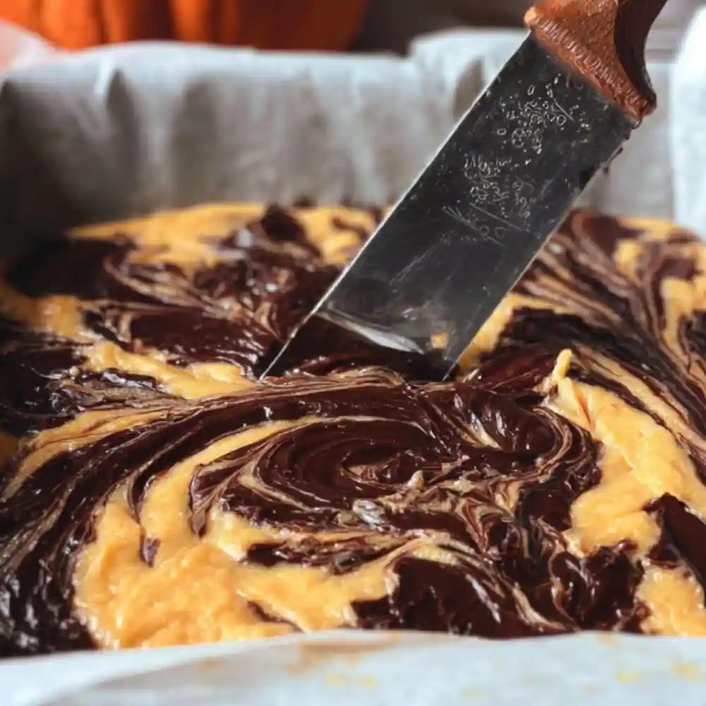 Swirled pumpkin brownie batter being marbled with a knife in a parchment-lined baking pan.