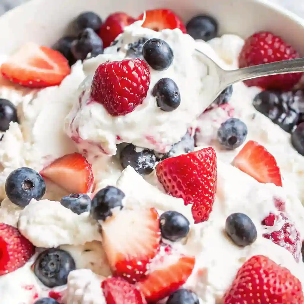 Spoonful of triple berry cheesecake fruit salad with strawberries, blueberries, and raspberries in a creamy mixture.