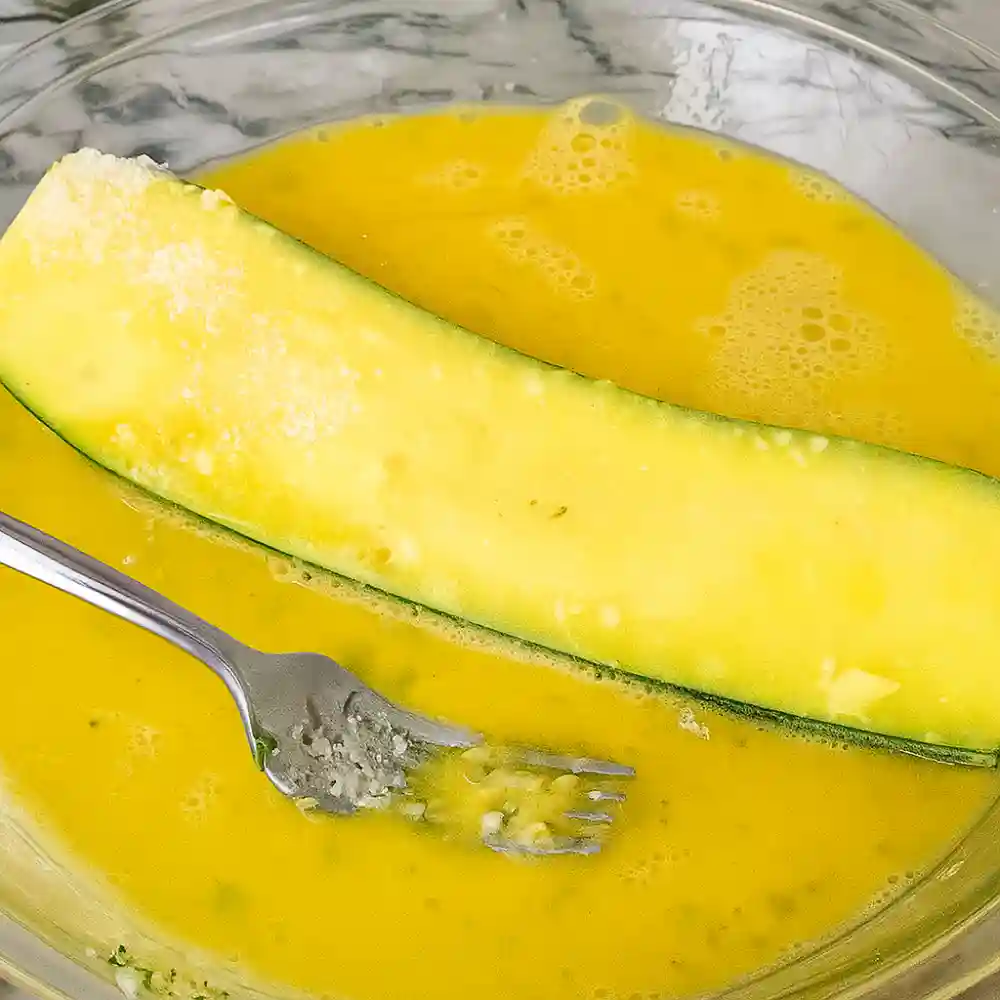 Zucchini slice dipped in beaten egg as part of breading process for Three Cheese Zucchini Parmesan