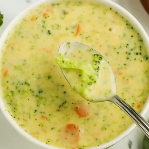 Spoonful of creamy high protein broccoli cheddar soup with broccoli and carrots in a white bowl.