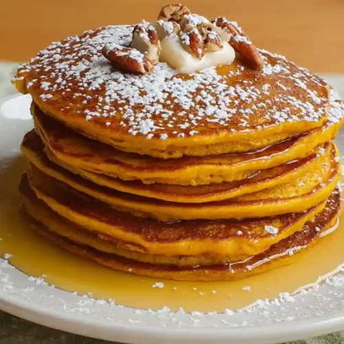 Stack of fluffy pumpkin pancakes topped with butter, pecans, powdered sugar, and maple syrup.