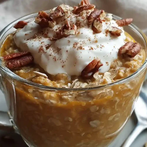 Glass cup filled with pumpkin pie overnight oats topped with whipped cream, pecans, and cinnamon.